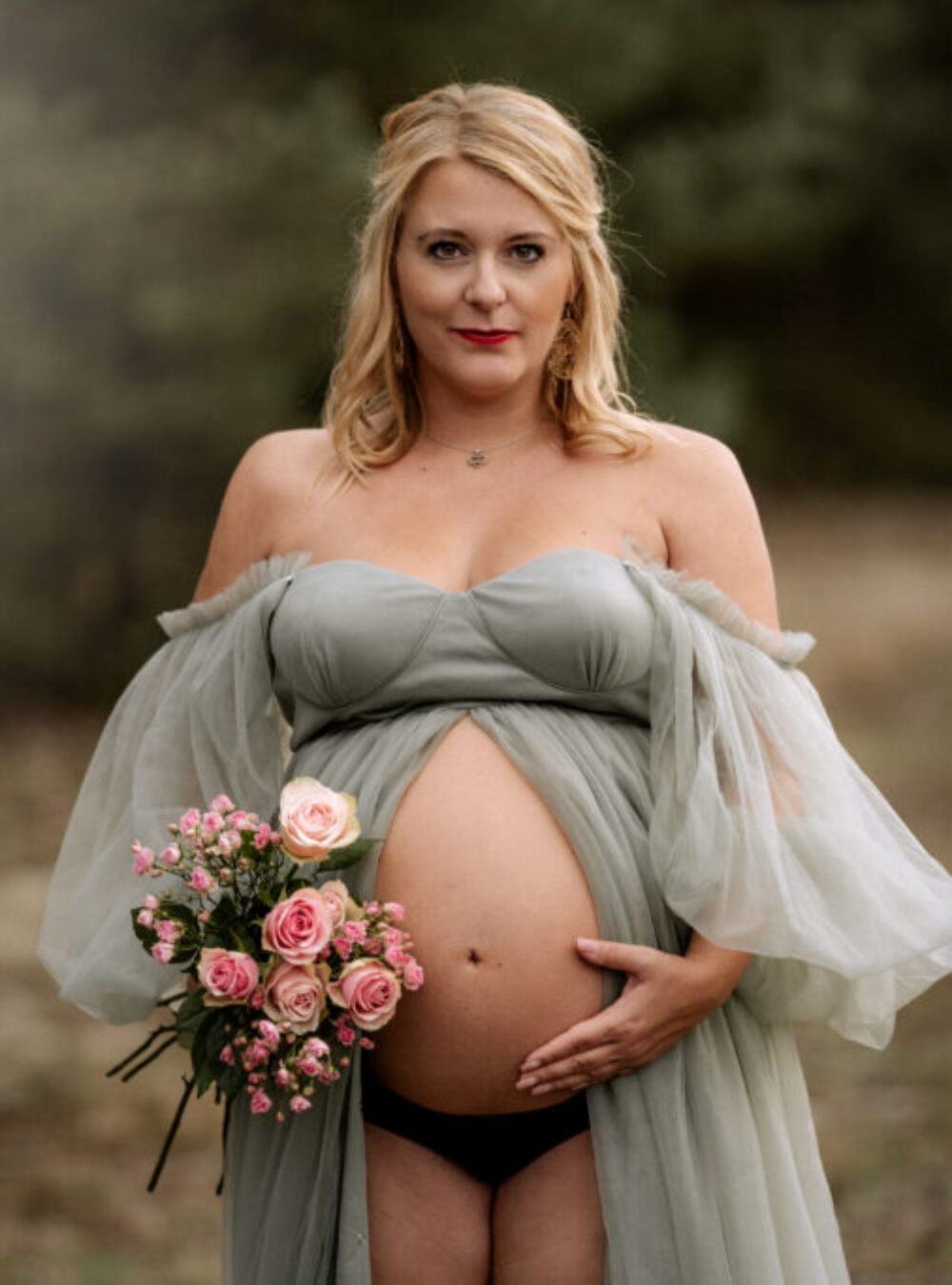 Hübsche, schwangere Frau im grünen Tüllkleid mit Rosen auf einer Waldlichtung.